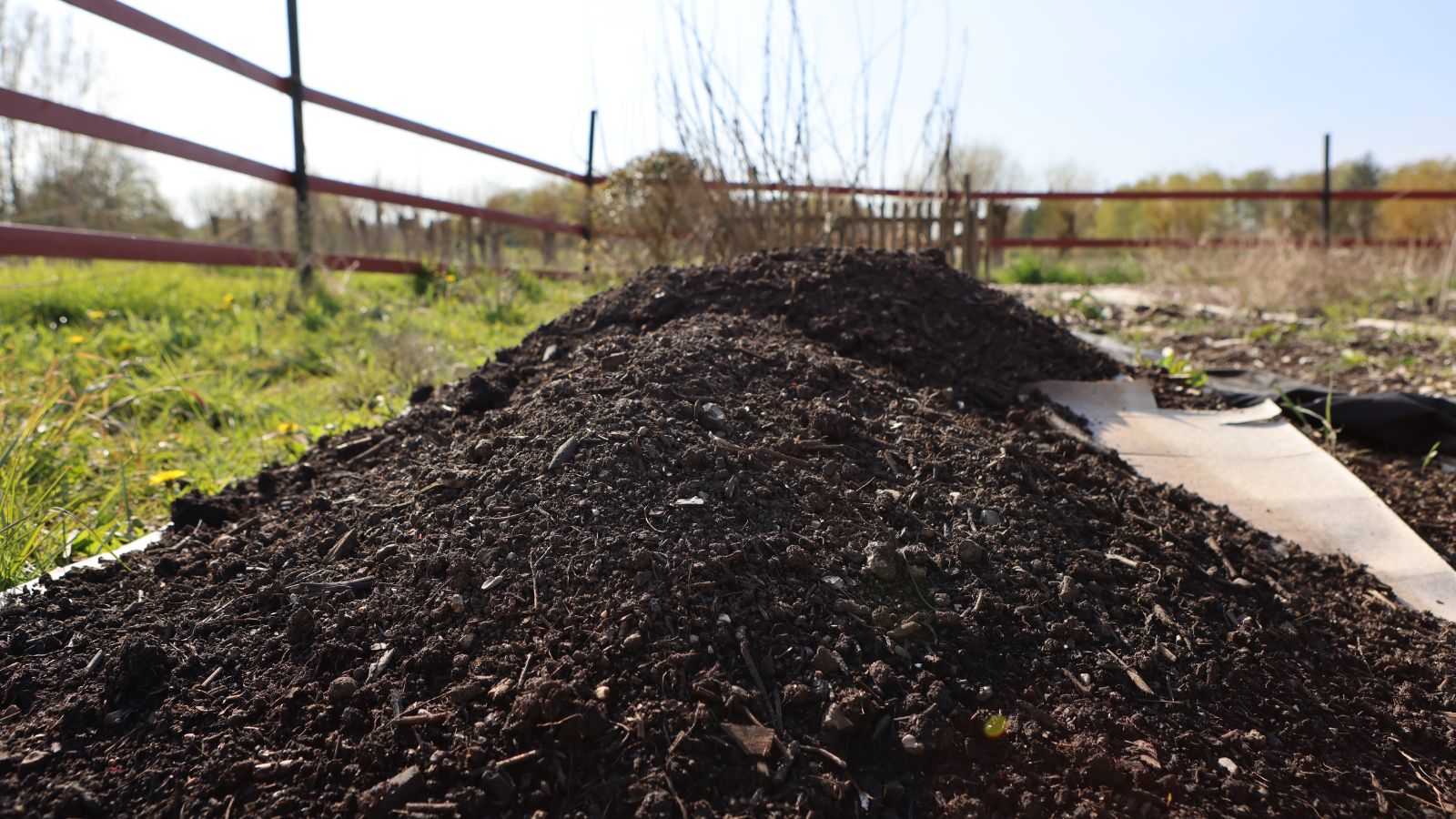 A pile of dark brown soil, being prepared, placed on mats for a garden bed, with fencing visible in the background